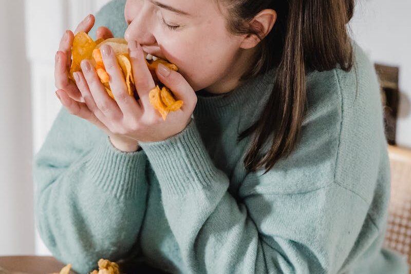 Hungry female in casual clothes eating crispy chips from hands while sitting at table with assorted junk food at home
