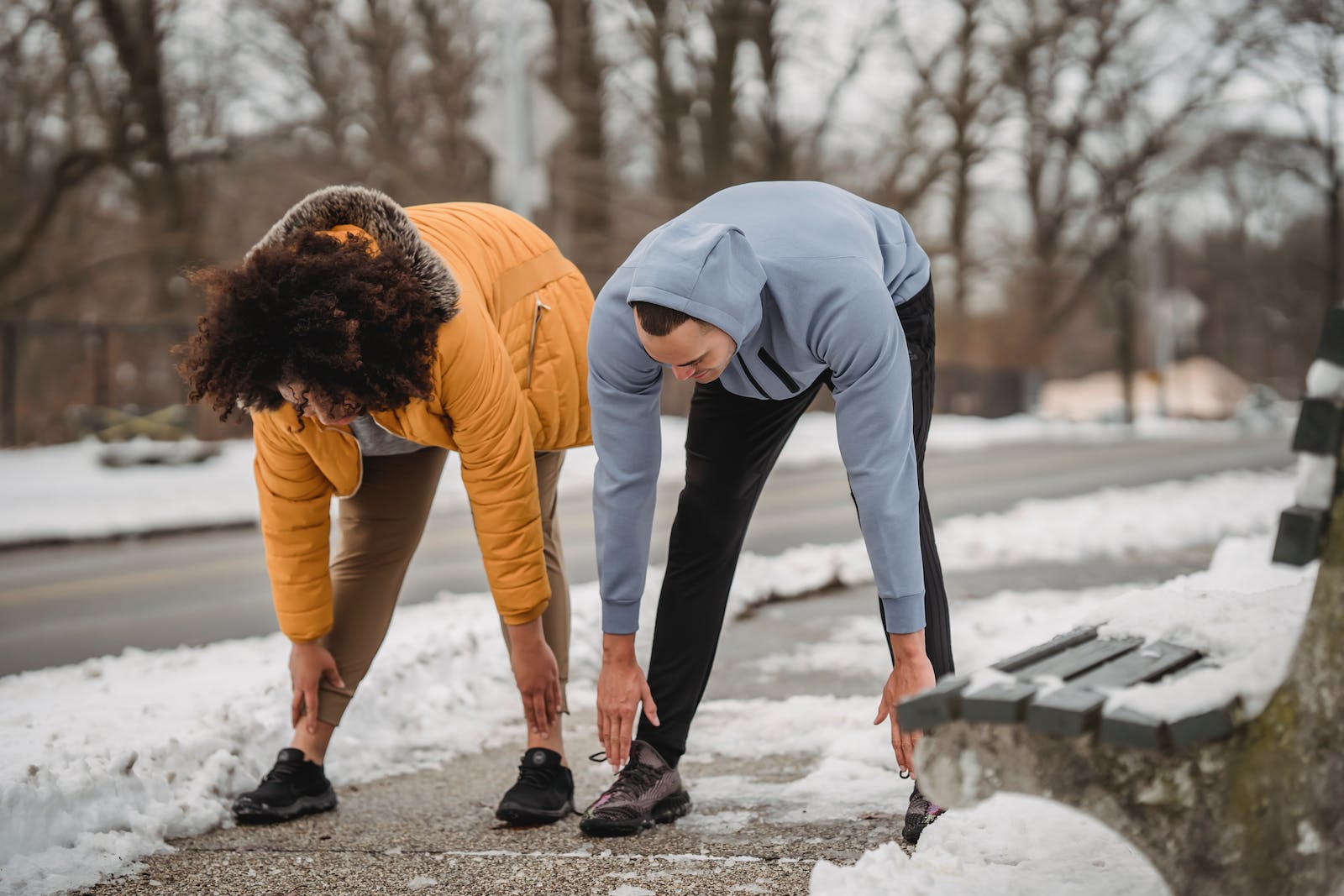 Full length anonymous fit male fitness trainer and plump trainer bending down while stretching body on snowy sidewalk in winter park
